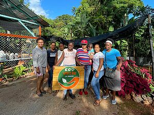 Das Foto zeigt sieben Erwachsene, die sich zu einem Gruppenbild formiert haben. Eine Frau trägt ein Kind auf dem Arm. In der Mitte hält eine Frau ein Holzschild mit dem Logo der Frauenkooperative „Eco Vivero La Sangría“ hoch.