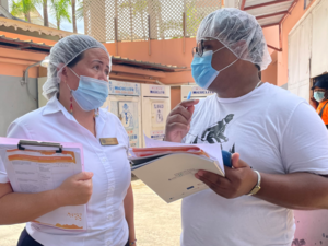Das Bild zeigt, wie eine Frau und ein Mann miteinander sprechen in der Dominikanischen Republik. Sie tragen eine medizinische Maske und eine Plastikhaube auf dem Kopf. In ihren Händen halten sie Dokumente.