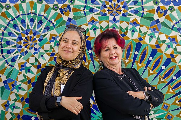 Zwei Frauen stehen Rücken an Rücken vor einer Mosaik-Wand. Sie haben die Arme vor der Brust verschränkt und lächeln in die Kamera.