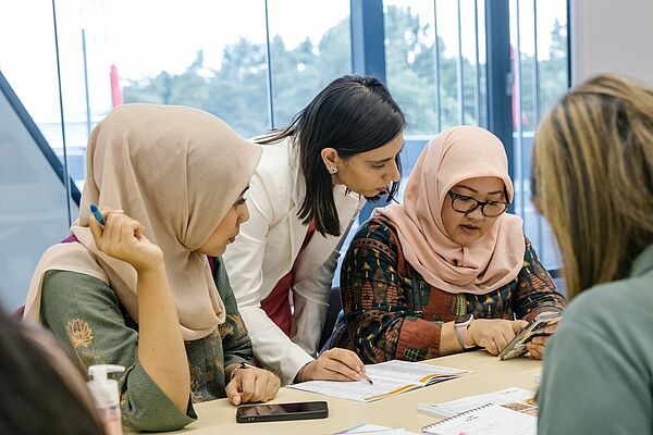 Drei Frauen sitzen an einem Tisch und arbeiten gemeinsam an Dokumenten, eine vierte Frau steht und zeigt auf die Papiere