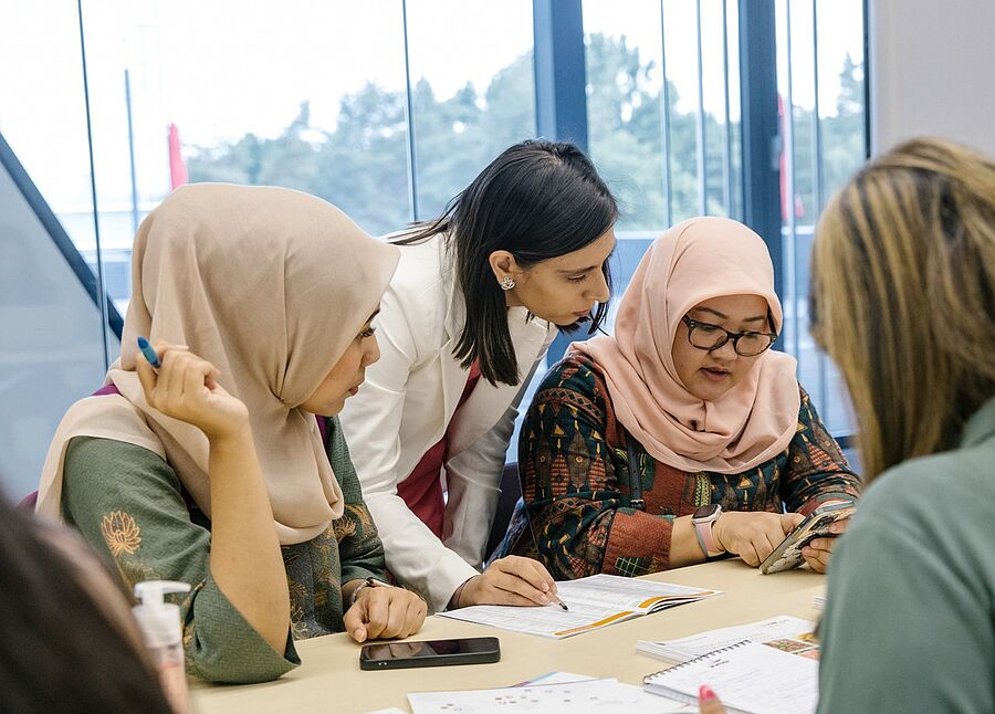 Drei Frauen sitzen an einem Tisch und arbeiten gemeinsam an Dokumenten, eine vierte Frau steht und zeigt auf die Papiere