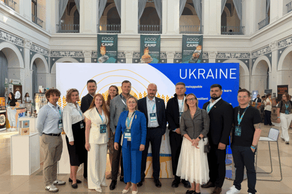 Eine Gruppe von Menschen posiert vor einer Messewand mit der Aufschrift ,Ukraine&lsquo; in einer gro&szlig;en Halle mit hohen Decken.