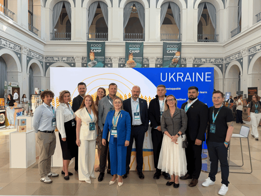 Eine Gruppe von Menschen posiert vor einer Messewand mit der Aufschrift ,Ukraine&lsquo; in einer gro&szlig;en Halle mit hohen Decken.