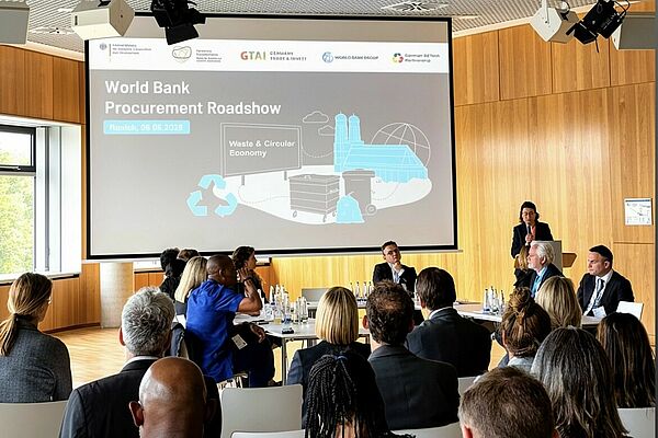 Im Vordergrund sitzen Teilnehmenden im Publikum, die auf eine U-f&ouml;rmige Tischordnung schauen, an der eine Gruppe von Experten diskutiert. Im Hintergrund projiziert eine Leinwand den Titel &bdquo;World Bank Procurement Roadshow&ldquo;.