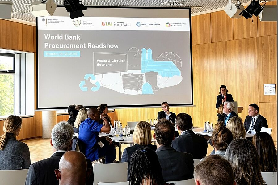 Im Vordergrund sitzen Teilnehmenden im Publikum, die auf eine U-f&ouml;rmige Tischordnung schauen, an der eine Gruppe von Experten diskutiert. Im Hintergrund projiziert eine Leinwand den Titel &bdquo;World Bank Procurement Roadshow&ldquo;.