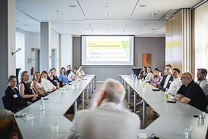 Ein Mann sitzt von hinten gesehen am Kopfende eines U-f&ouml;rmigen Konferenztisches, w&auml;hrend im Hintergrund eine Pr&auml;sentation mit dem Titel &bdquo;Roundtable. Tourismus, Verantwortung, Entwicklung&ldquo; an die Wand projiziert wird.