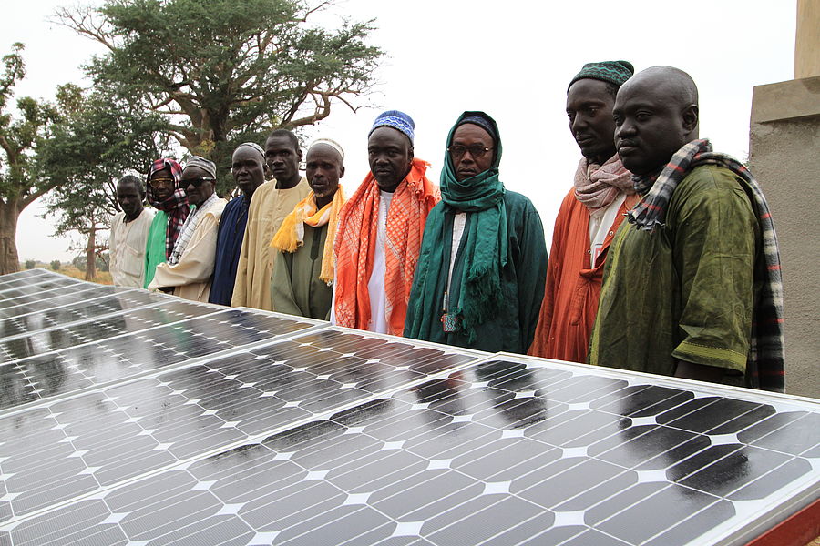 Auf einem Feld im Senegal stehen Menschen hinter Photovoltaikplatten. Im Hintergrund sind Bäume.